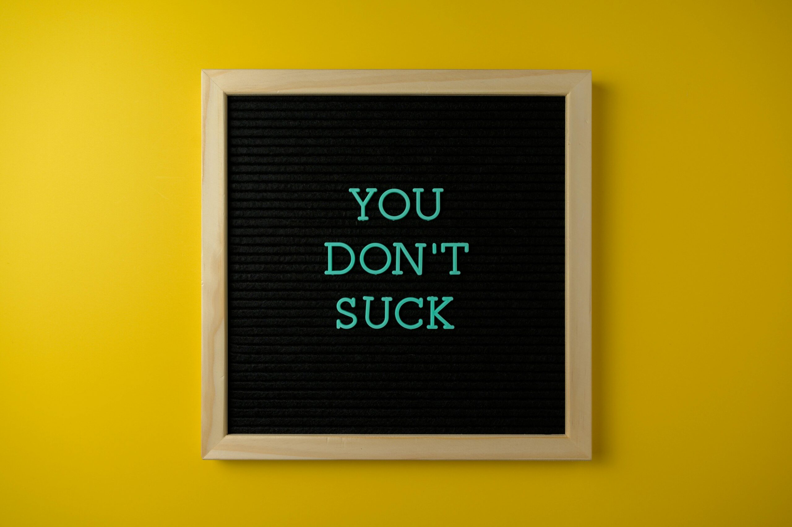 A black felt letter board with a light wooden frame displays the message "YOU DON'T SUCK" in teal letters, centered against a bright yellow background. A motivational reminder for what to do when you don't feel like it.
