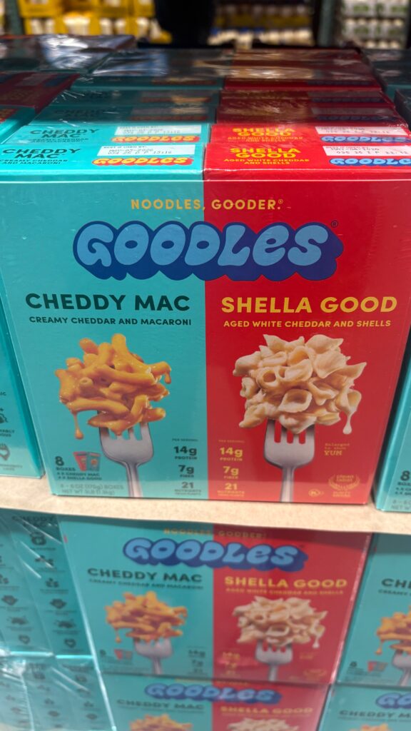 Boxes of Goodles brand mac and cheese on a store shelf, featuring two flavors: "Cheddy Mac" (creamy cheddar and macaroni) in a turquoise box, and "Shella Good" (aged white cheddar and shells) in a red box. Both highlight 14g protein and 21 nutrients.